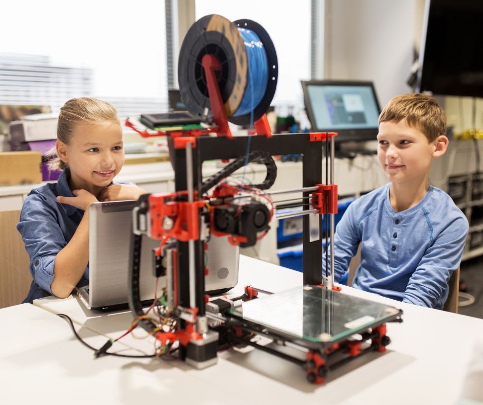 Two children sitting at a table. One has a laptop open in front of them. Both are watching a 3D printer.